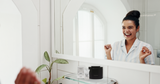 A woman in a white shirt smiling at a mirror, delighted with a smile probably maintained via a retainer replacement subscription.