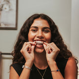 A smiling woman with long dark curly hair is shown from the chest up, wearing a clear dental aligner tray and pulling it slightly outward with both hands. She is wearing a black sleeveless top, silver bracelets, and a gold necklace in an indoor setting with a light gray wall and framed picture in the background.