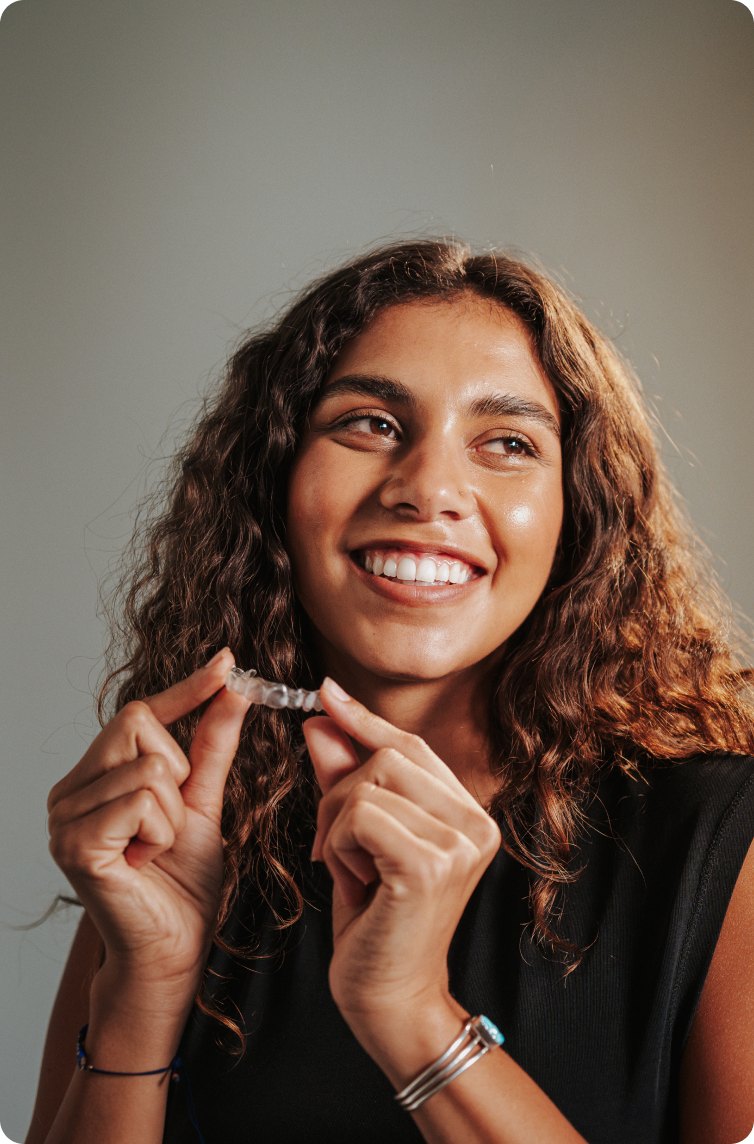 A smiling woman holding a clear aligner up to her teeth